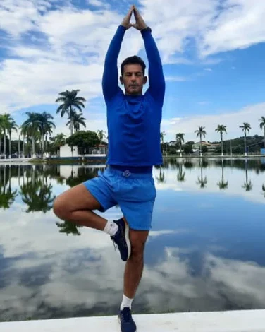 Roberto na postura da árvore, de braços erguidos, à beira de um lago com palmeiras refletidas na água